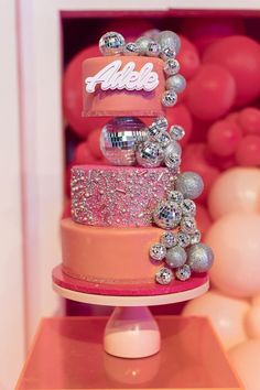 a three tiered cake with pink and silver decorations on the top, surrounded by balloons