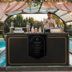 a man standing behind a bar with drinks on it in front of a swimming pool