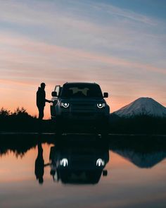 a man standing next to a parked car in front of a body of water at sunset