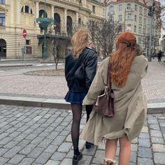 two women are walking down the sidewalk in front of some buildings and one is wearing a brown coat