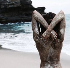 a woman covered in sand sitting on the beach with her back to the camera and hands behind her head