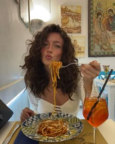 a woman sitting at a table eating spaghetti from a plate with a drink in front of her