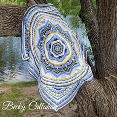 a blue and yellow crocheted blanket hanging from a tree next to a body of water