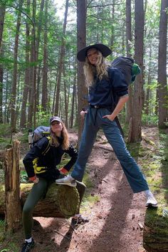 two people standing in the woods with backpacks on their back and one person sitting on a log