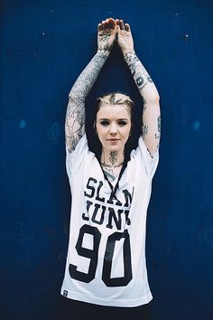 a woman with tattoos standing in front of a blue wall holding her hands above her head