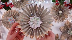 two hands are holding some paper flowers on a white surface with christmas decorations in the background