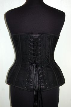 "This is our standard hipgore overbust corset, made in an embroidered raw silk. It's a black on black floral & foliage design. It is also back laced with a matching black double satin quarter inch wide black ribbon and front busk fastening, hand made matching silk bias binding top and bottom to match the boning casings and main body. Comes compete with a loose style modesty panel. The silk is backed with woven vilene, which then has a British herring bone cotton coutil lining. All lovingly c