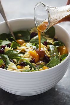 a person pouring dressing into a bowl of pasta salad with spinach and oranges