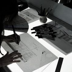 a woman is drawing on some paper at a table with pens and pencils in front of her