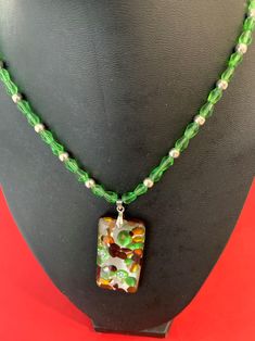 Lampwork Rectangular Glass Pendant Necklace measures approximately 20" from clasp to closure. The pendant dangles 1 1/4"  and is 1" wide. The depth is 1/4". It can be lengthened or shortened at no extra charge. The lampwork pendant is vibrantly colored in  green, topaz, raspberry on a gray background.  The reverse side of the pendant is raspberry. The pendant contains a bale that is 1/4" in length and connects it to a necklace of 42 faceted green teardrop beads in pairs of two, measuring each 1/4" in size and is separated by 24 smooth silver spacer beads.   There are two crimp cover beads on each side of the necklace near the closing.  Finally, the necklace is completed with a silver heart toggle clasp measuring 3/4"  This necklace is an eye-catcher and makes a beautiful gift.  It will dre Unique Green Glass Necklace, Unique Glass Necklace Pendant Folksy, Artisan Hand-strung Glass Necklaces, Elegant Green Murano Glass Necklace, Green Topaz, Lampwork Pendant, Lampwork Necklace, Glass Pendant Necklace, Teardrop Beads