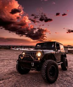 a jeep parked in the middle of a dirt field at sunset with clouds above it