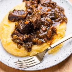 a white plate topped with meat and grits next to a fork on top of a wooden table