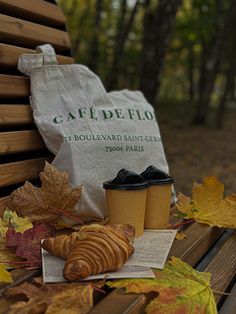 two coffee cups sitting on top of a wooden bench next to leaves and a bag