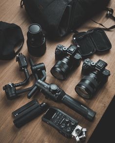 many different cameras and accessories on a table with a camera bag in the back ground