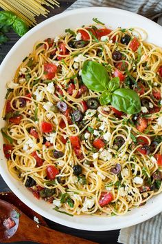 a white bowl filled with pasta, olives and spinach on top of a wooden cutting board