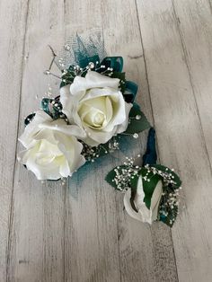 two white roses with green leaves and pearls on them sitting on a wooden floor next to each other