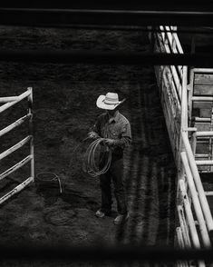 a man with a cowboy hat standing next to a fence holding a lasso in his hand