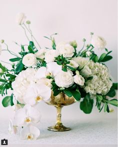 white flowers and greenery in a gold vase on a table with a white background