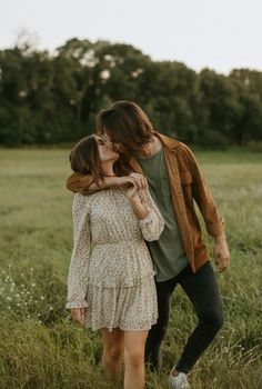 two people are walking through the tall grass in an open field, one is kissing the other's cheek