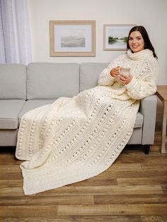 a woman sitting on a couch holding a coffee cup and wearing a white crocheted blanket