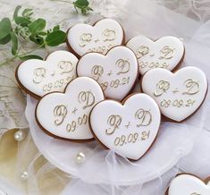 cookies decorated with initials and hearts on a white plate next to greenery, pearls and lace