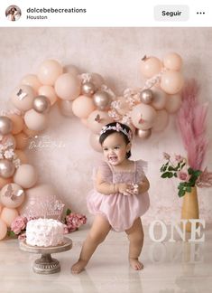 a baby girl standing in front of a cake with balloons and flowers on the wall