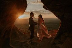 a man and woman standing in front of a cave with the sun setting behind them