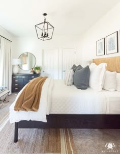 a bed with white sheets and pillows in a bedroom next to a dresser, mirror and chandelier