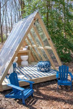 a small house made out of wood and glass in the woods with blue chairs around it