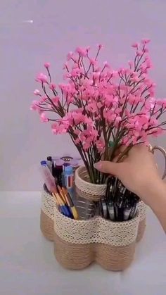 a hand holding a cup filled with pens and pencils next to a flower pot