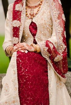 a woman in a red and white wedding outfit sitting on a stone wall with her hands clasped to her chest