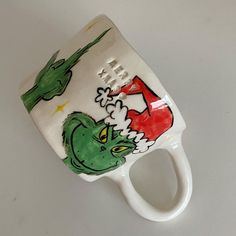 a ceramic mug with an image of the grinch on it's side, sitting on a white surface