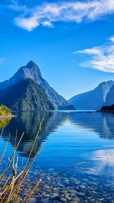 the mountains are reflected in the still water on the lake's edge, while grass and rocks stand out against the blue sky