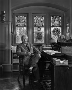 an old black and white photo of a man sitting at a desk in front of stained glass windows