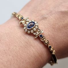 "A beautiful vintage bangle bracelet, the face featuring an oval cluster consisting of a blue sapphire surrounded by white pearls, the shoulders of the bangle with leaf details and four further sapphires. The bangle is hinged, with a push-in tongue clasp and safety chain. Hallmarked as 9ct gold, dated London 1990. Maker's mark \"DWT\". The inner circumference measures approximately 170mm. The central cluster measures approximately 11.8mm (L) x 10mm (W). The approximate weight is 12.0 grams. COND Gold Vintage Bracelet, Vintage Bangle Bracelets, Pearl Bangle, Vintage Sapphire, Vintage Bangles, Safety Chain, Vintage Bracelet