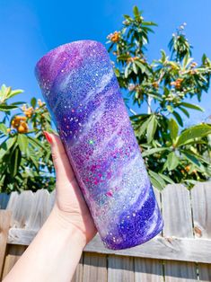 a hand holding up a purple and blue tube with glitter on it in front of a fence