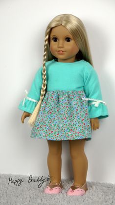 a doll with long blonde hair wearing a blue dress and pink shoes, standing against a white wall