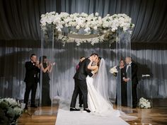 a bride and groom kissing under an arch decorated with white flowers at their wedding ceremony