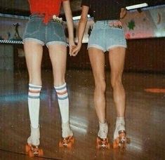 two women in short shorts and socks standing on a wooden floor with roller skates