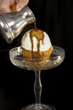 someone pouring caramel sauce on an ice cream sundae in a glass dish with a silver shaker