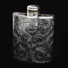 a silver flask with an ornate design on the front and side, sitting against a black background