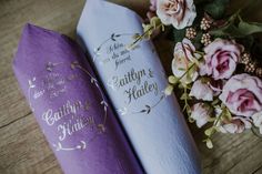 two purple wedding paper napkins sitting on top of a wooden table next to flowers