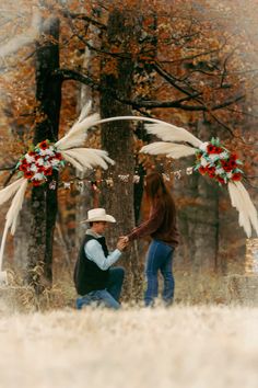 a man kneeling down next to a woman in front of a tree with white feathers