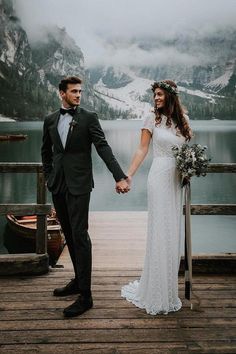 a bride and groom holding hands on a dock in front of a lake with mountains