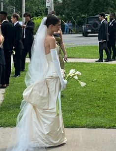 the bride is walking down the sidewalk with her bouquet in hand and other people behind her