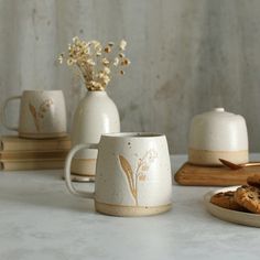 three white mugs and two cookies are on the table next to each other with flowers in them