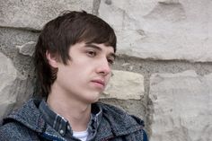 a young man leaning against a stone wall with his head turned to the side and looking off into the distance