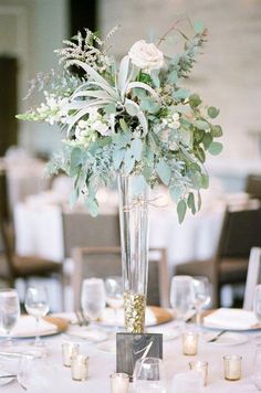 a tall vase filled with flowers and greenery on top of a white table cloth