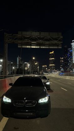 two cars parked on the side of a road at night with city lights in the background