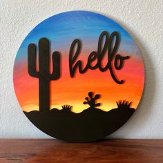 a wooden sign that says hello, with a cactus in the foreground and an orange sky behind it